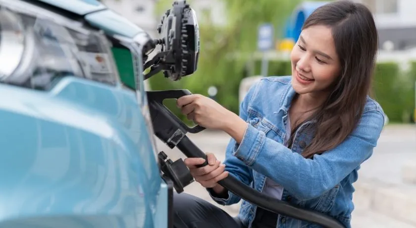 一位女士在公寓大樓外享用咖啡並檢查手機，同時她的電動車正在充電